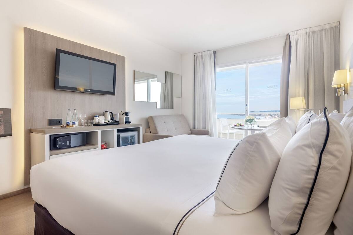 A hotel room with a large bed, a TV mounted on the wall, a desk with a microwave, and a view of the sea.
