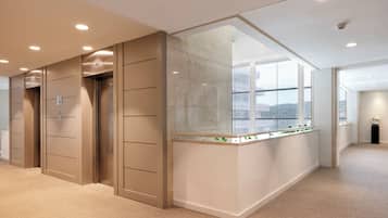 Un área de ascensor moderna con paredes color beige, un panel de vidrio y vistas de un edificio a través de grandes ventanales.