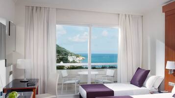Habitación de hotel con cama, escritorio, silla y mesa. Amplia ventana con vista al mar y un edificio.