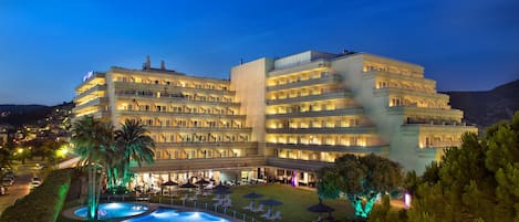 A multi-story hotel with a pool area, outdoor seating, and a well-lit facade.