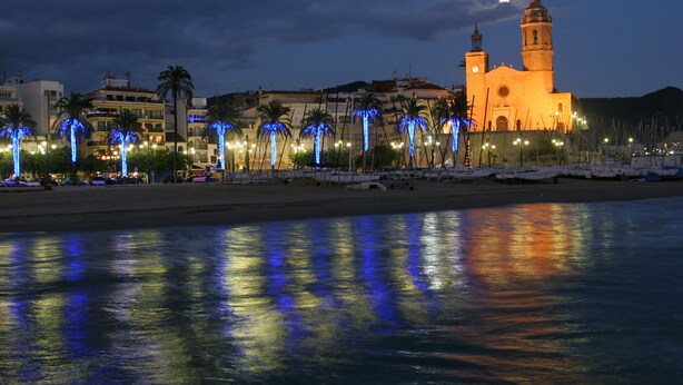A waterfront area with a prominent building featuring two towers, palm trees with blue lights, and a calm body of water reflecting the lights. - Melia Sitges (Sitges)