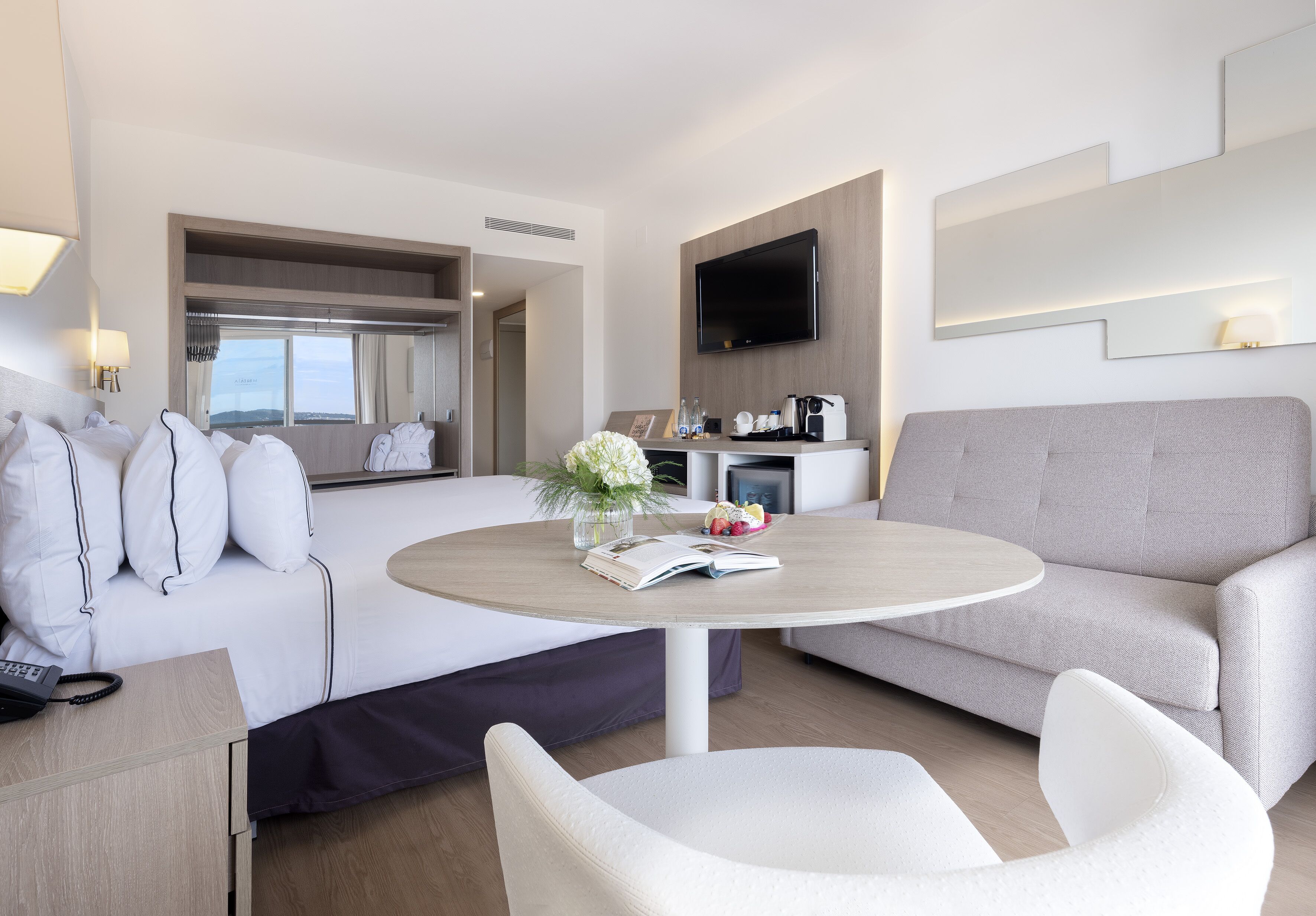 Una habitación de hotel moderna con una cama, un sofá, una mesa redonda con un libro y flores, un televisor y una cafetera.