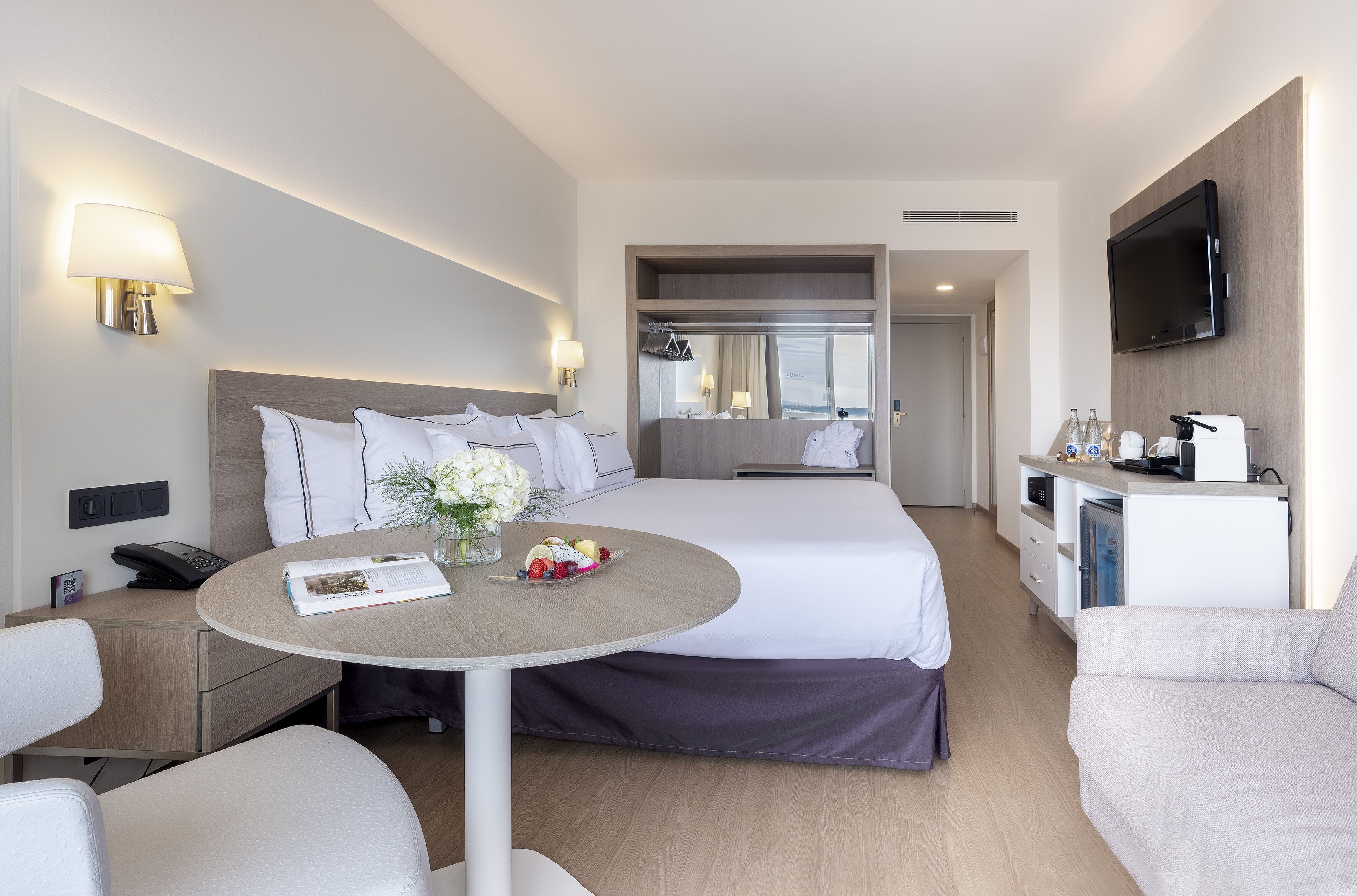 A modern hotel room with a bed, a dining table with a vase of flowers, a sofa, a TV, and a minibar.