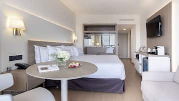 A modern hotel room with a bed, a dining table with a vase of flowers, a sofa, a TV, and a minibar.