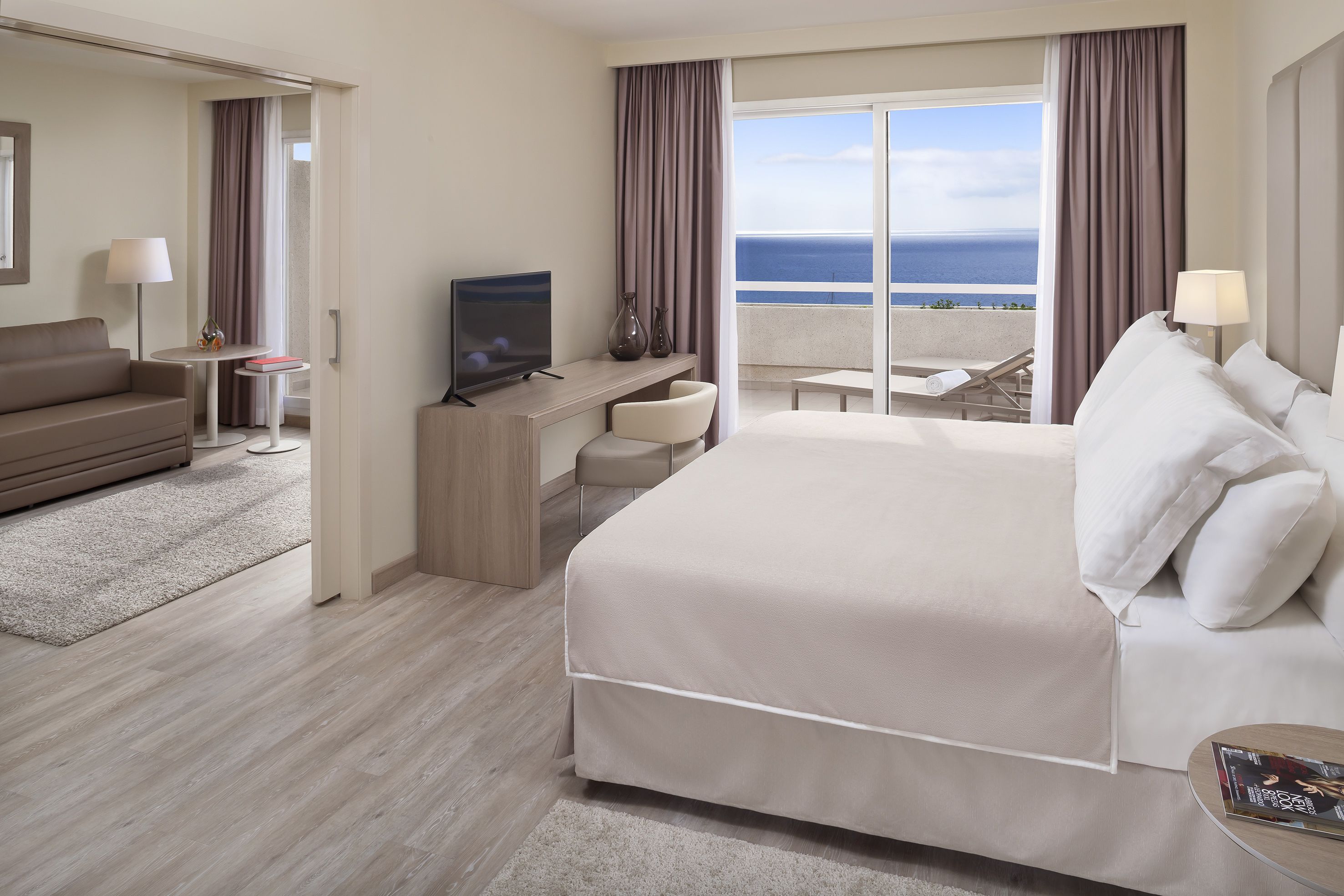 Ein Hotelzimmer mit einem großen Bett, einem Schreibtisch mit Fernseher, einer Sitzecke und einem Balkon mit Meerblick.