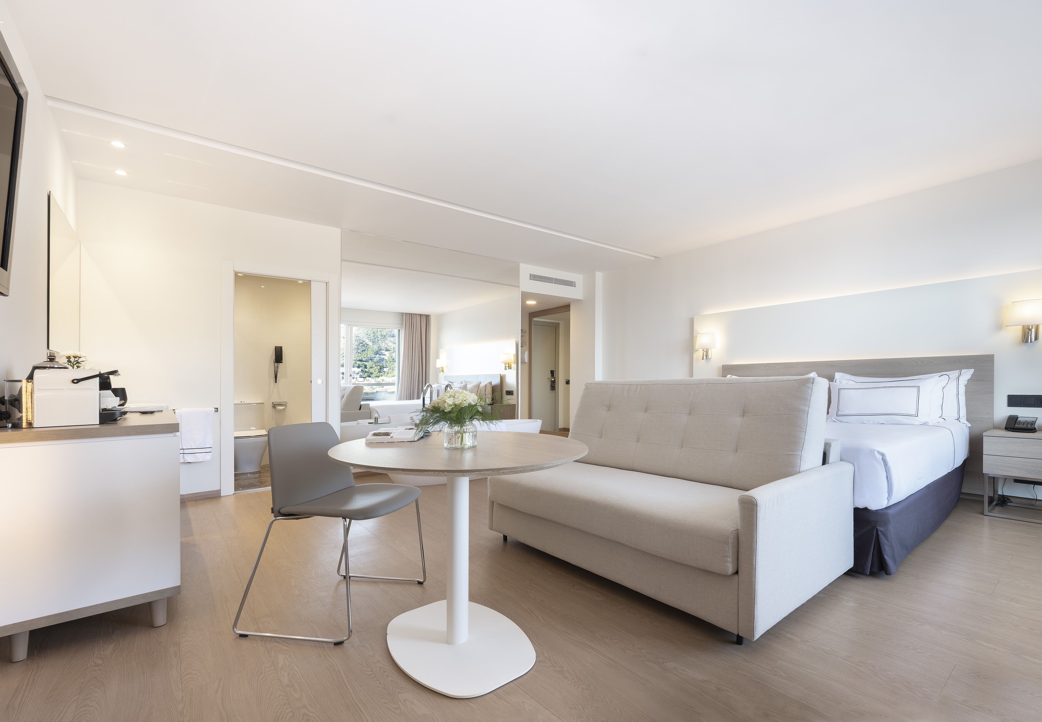 A modern hotel room with a round dining table, a sofa, a bed, and a desk with a chair.