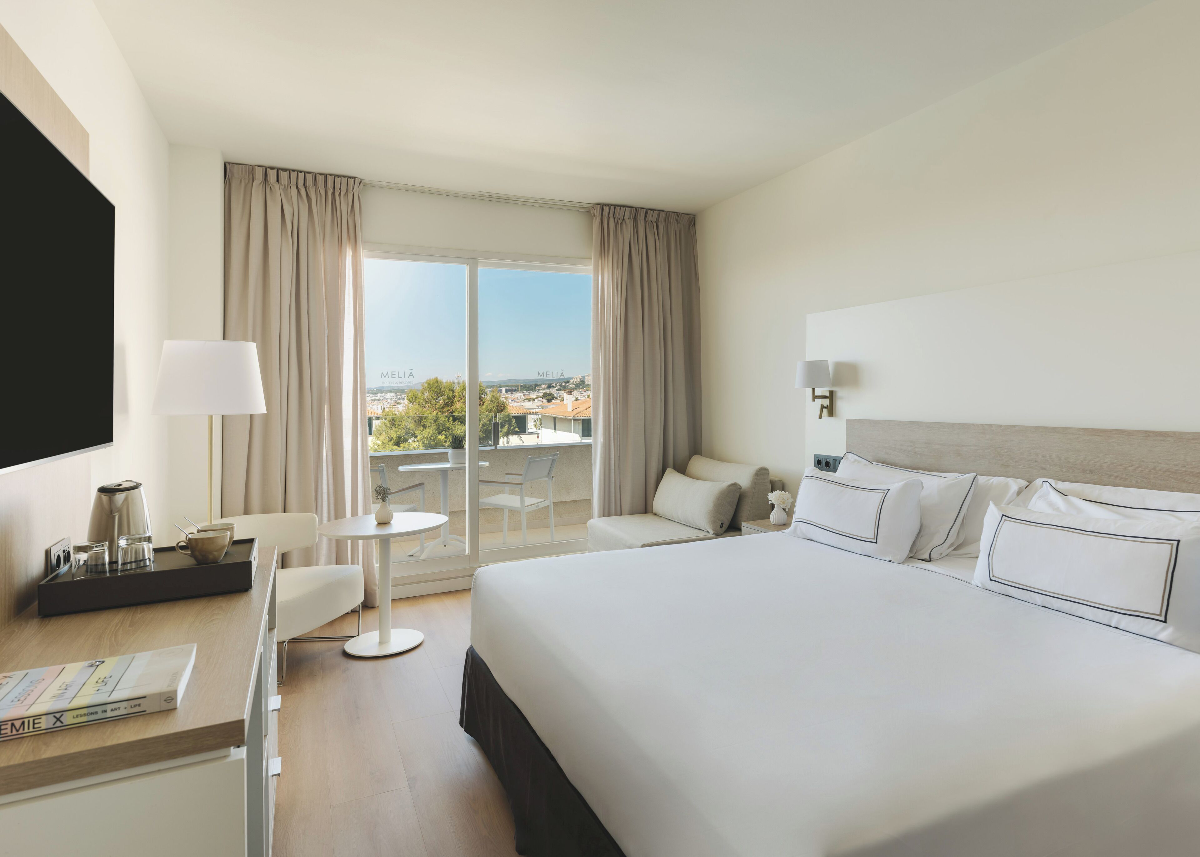 A hotel room with a large bed, a desk with a coffee maker, a chair, and a view of the city through large windows.