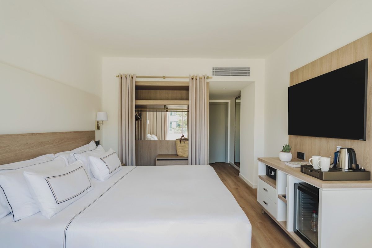 Habitación de hotel con una cama grande, televisión, minibar y vistas al pasillo.