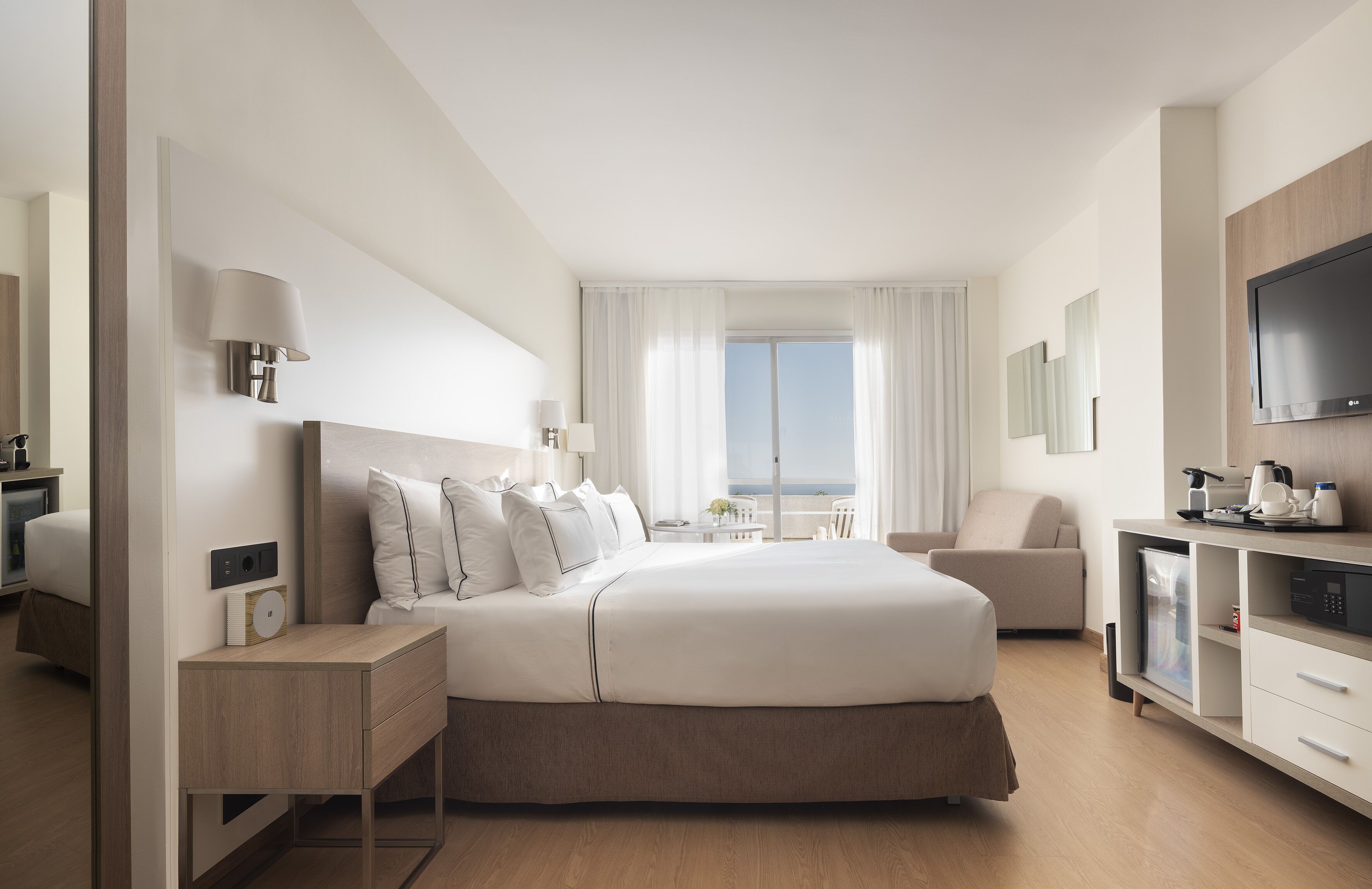 A modern hotel room with a large bed, bedside tables, a TV, and a view of the sea.