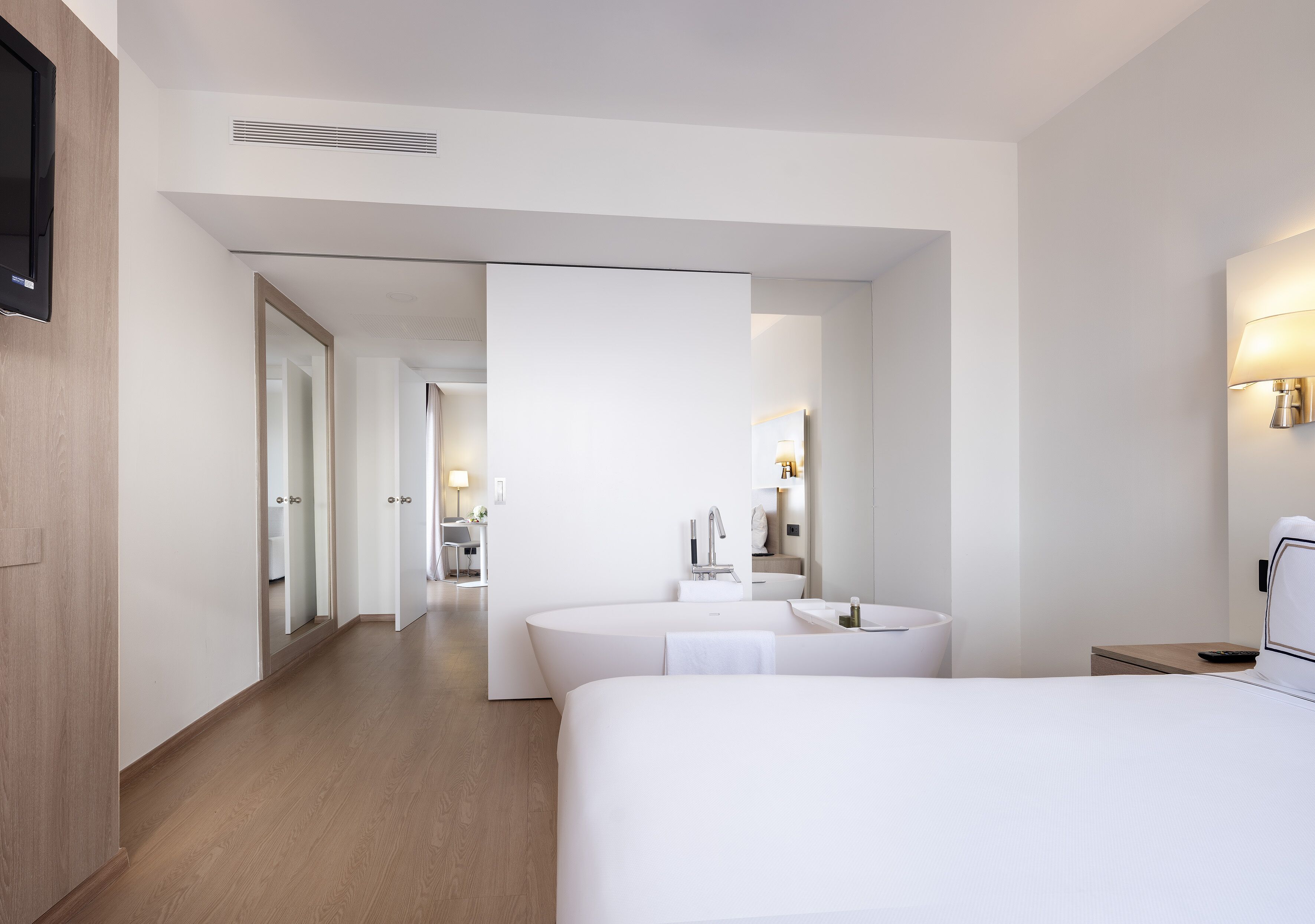 A modern hotel room with a large bed, a sitting area, a bathroom with a bathtub, and a hallway leading to other rooms.