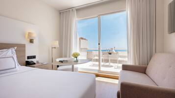 Een hotelkamer met een bed, een bureau, een stoel, een tafeltje met een bloevas en een balkon met uitzicht op zee.