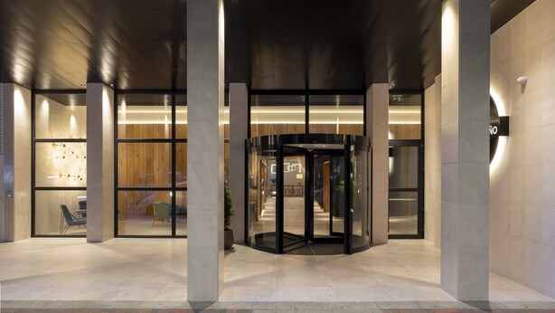 Una entrada moderna de edificio con puerta giratoria, amplios ventanales de vidrio y zona pavimentada con ladrillos.