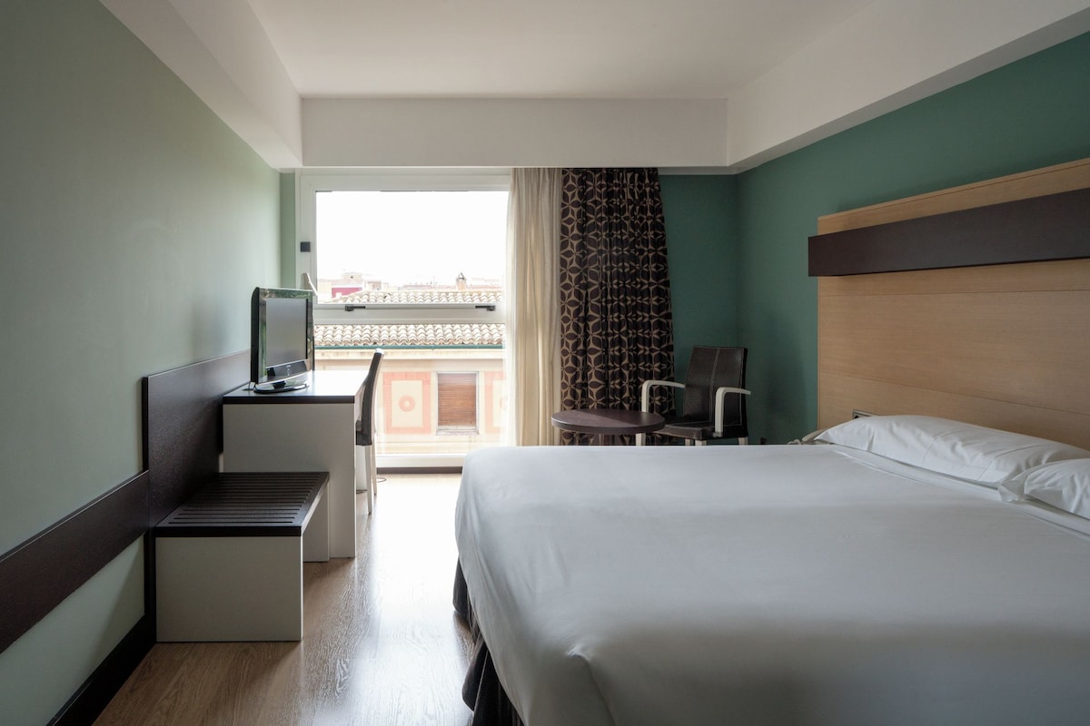 A hotel room with a bed, a desk with a computer, a chair, and a view of the city through a window.