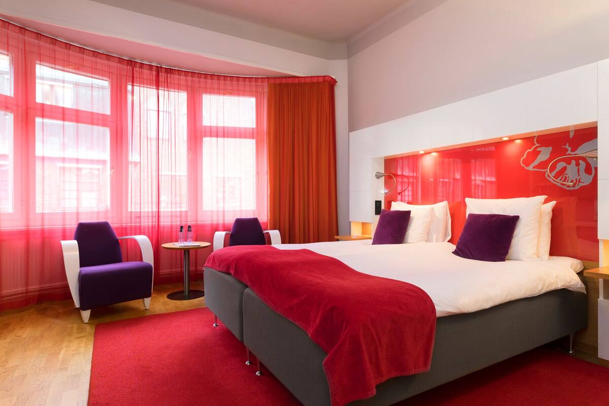 A modern hotel room with a large bed, two armchairs, a small table, and a red carpet.