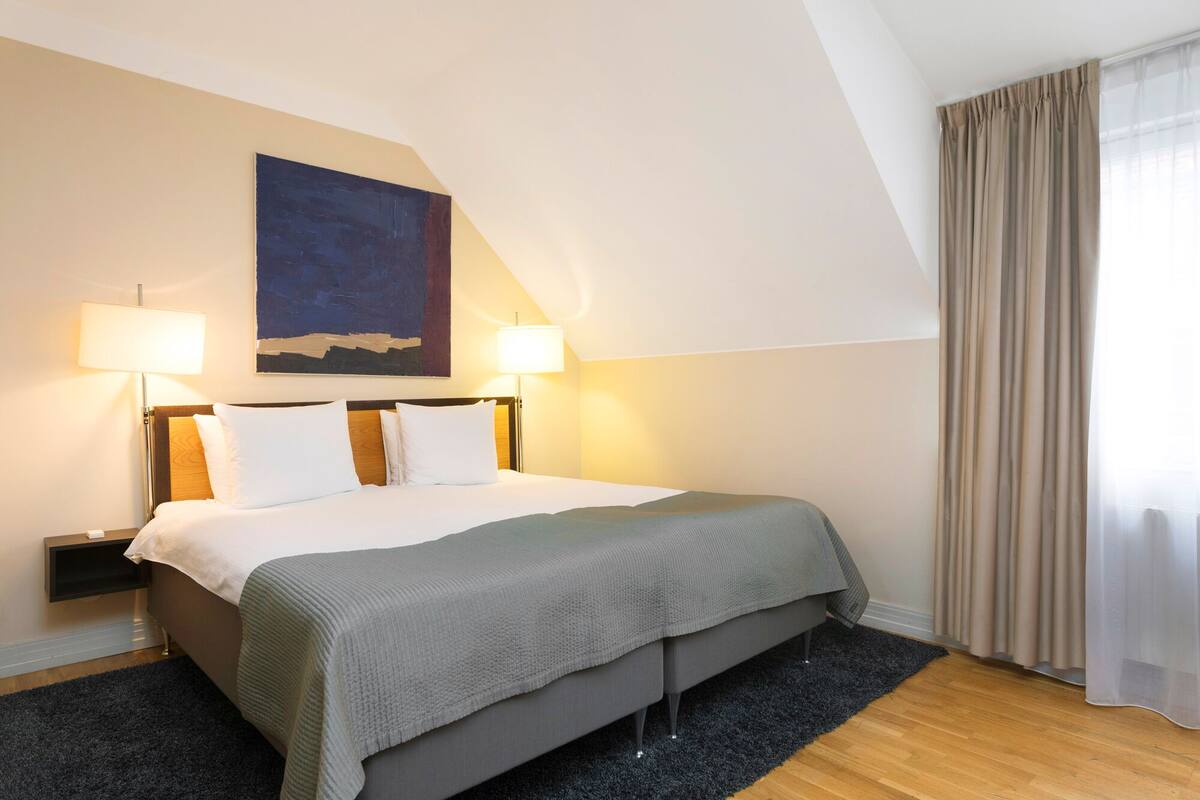 A hotel room with a bed, two bedside lamps, a nightstand, a dark rug, and a window with curtains.
