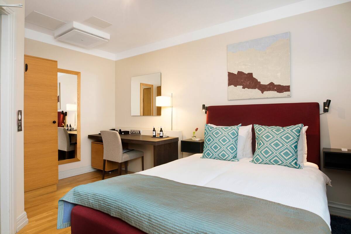 A hotel room with a bed, a desk, a chair, a mirror, a wall-mounted lamp, and a painting.