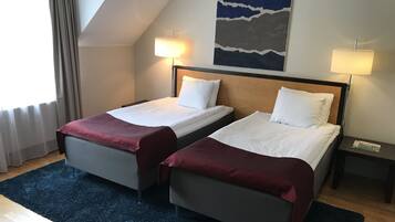 Habitación de hotel con dos camas individuales, un ventanal grande con cortinas, un cuadro en la pared y una mesita de noche con lámpara.