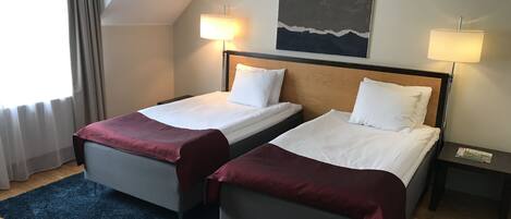 Habitación de hotel con dos camas individuales, un ventanal grande con cortinas, un cuadro en la pared y una mesita de noche con lámpara.