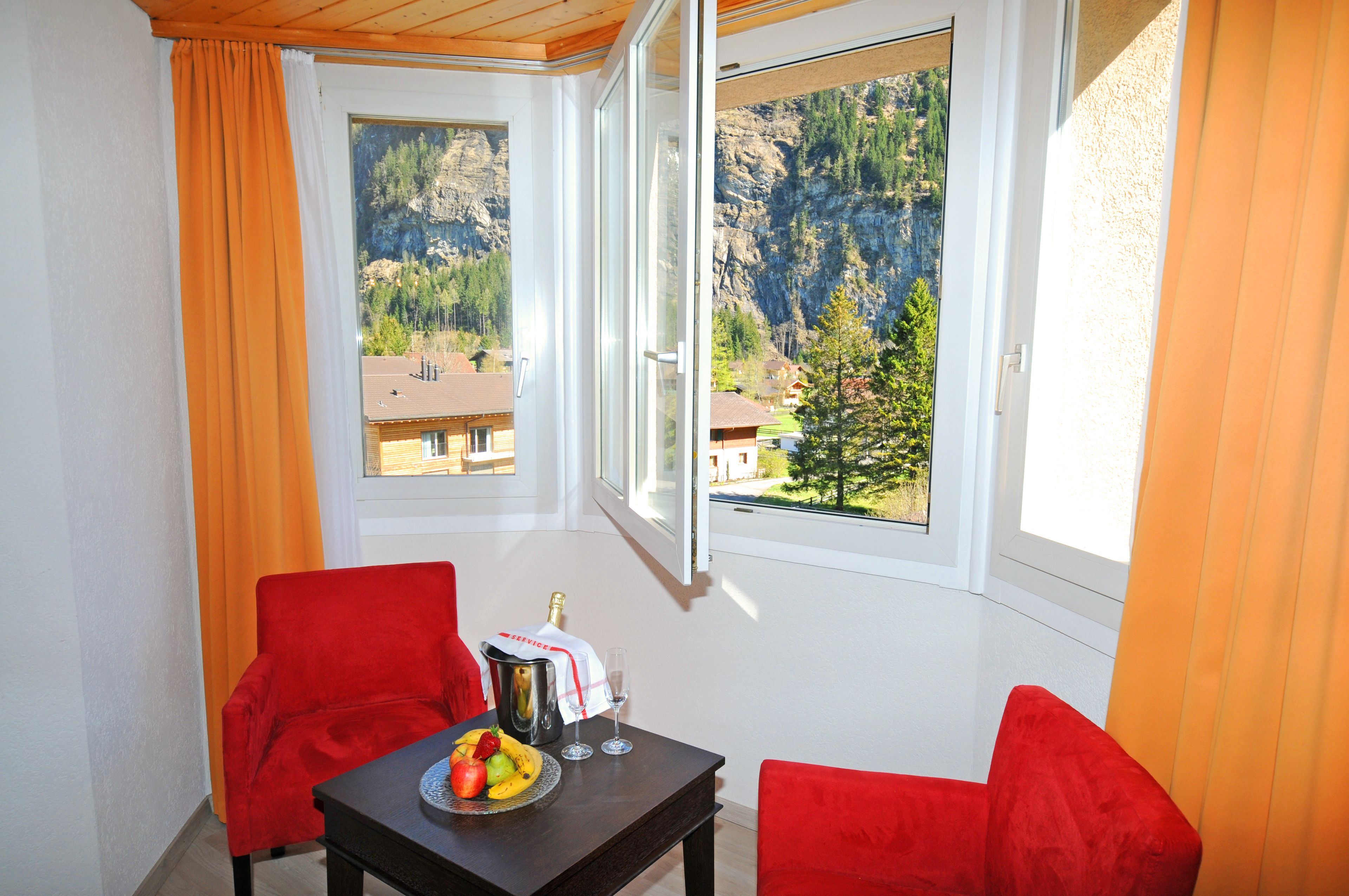 A balcony with two red chairs, a table with fruit and a bottle of champagne, and a view of a mountainous landscape.