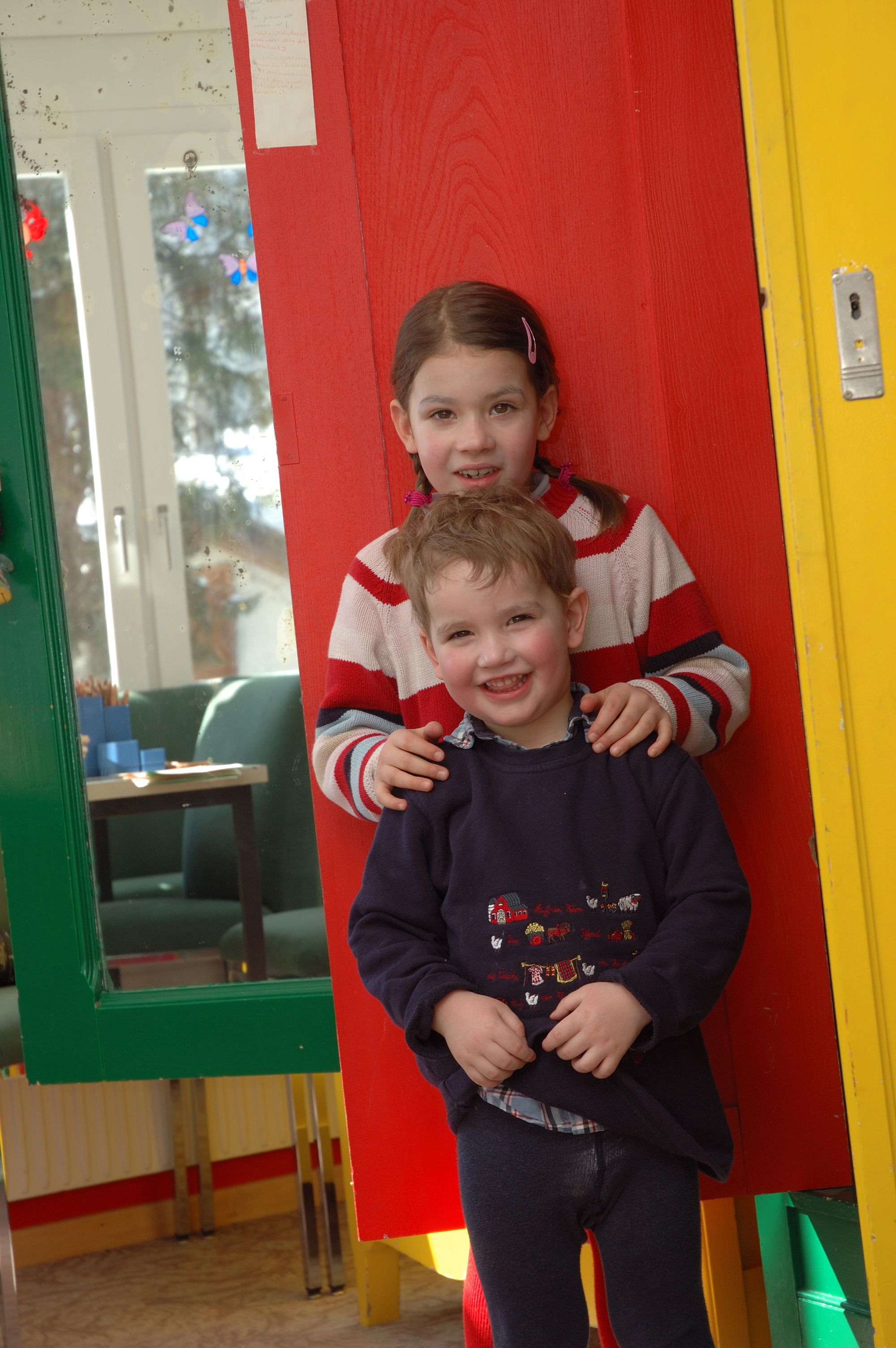 Two children standing in a room with a red and yellow door, a green table, and a window with decorations.