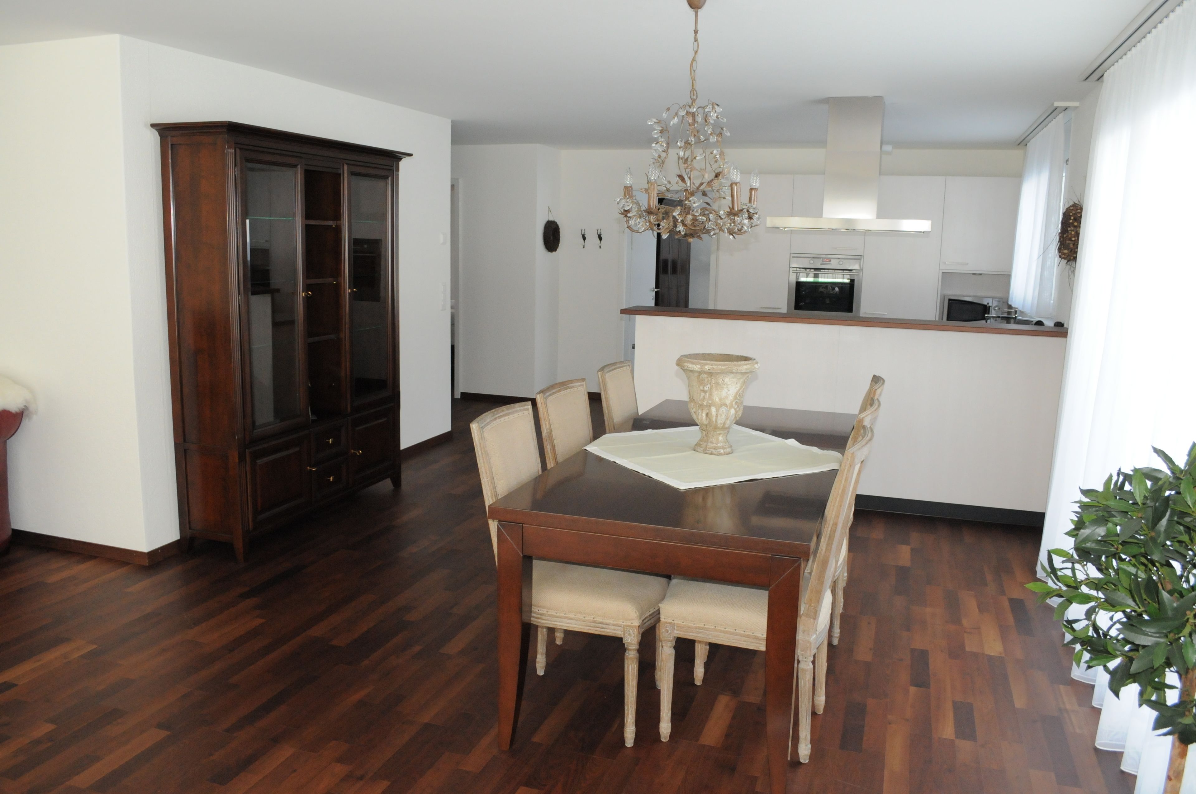 A dining area with a wooden table and chairs, a chandelier, and a kitchen in the background.