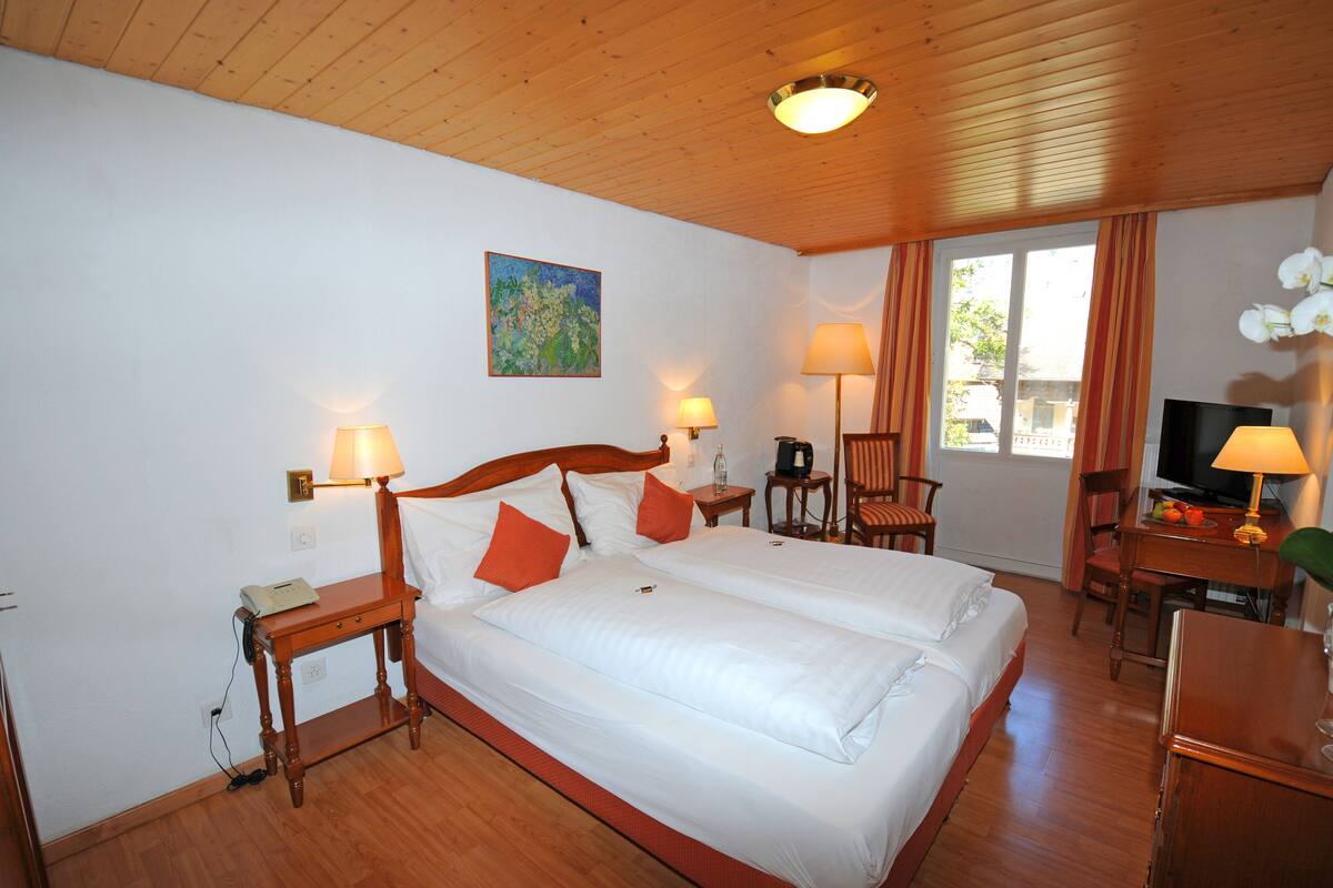 Ein Hotelzimmer mit zwei Betten, Holzböden, einem Schreibtisch mit Stuhl, einem Fernseher und einem Fenster mit Vorhängen.