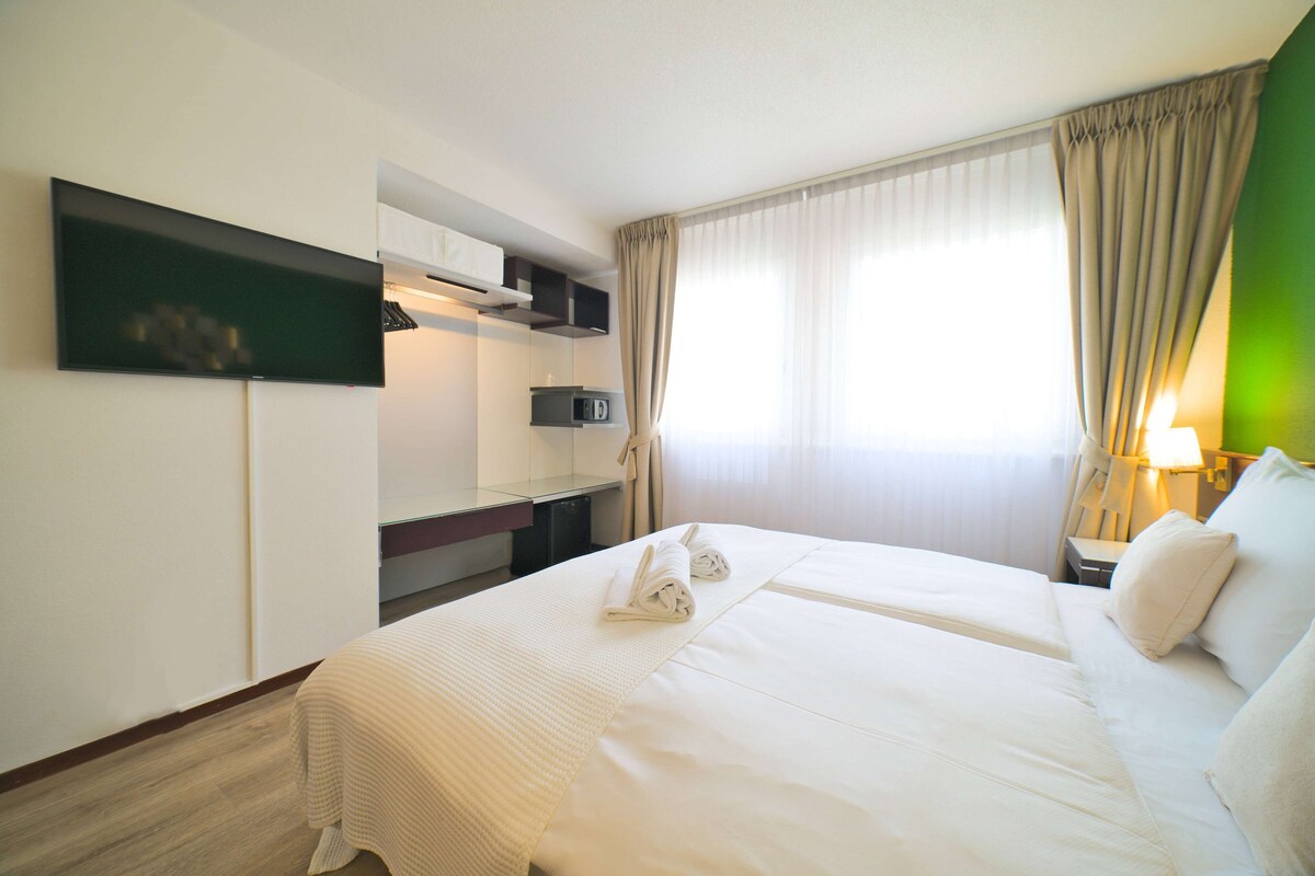 A hotel room with a large bed, a flat-screen TV mounted on the wall, a desk with a computer, and a window with curtains.