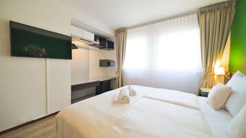A hotel room with a large bed, a flat-screen TV mounted on the wall, a desk with a computer, and a window with curtains.
