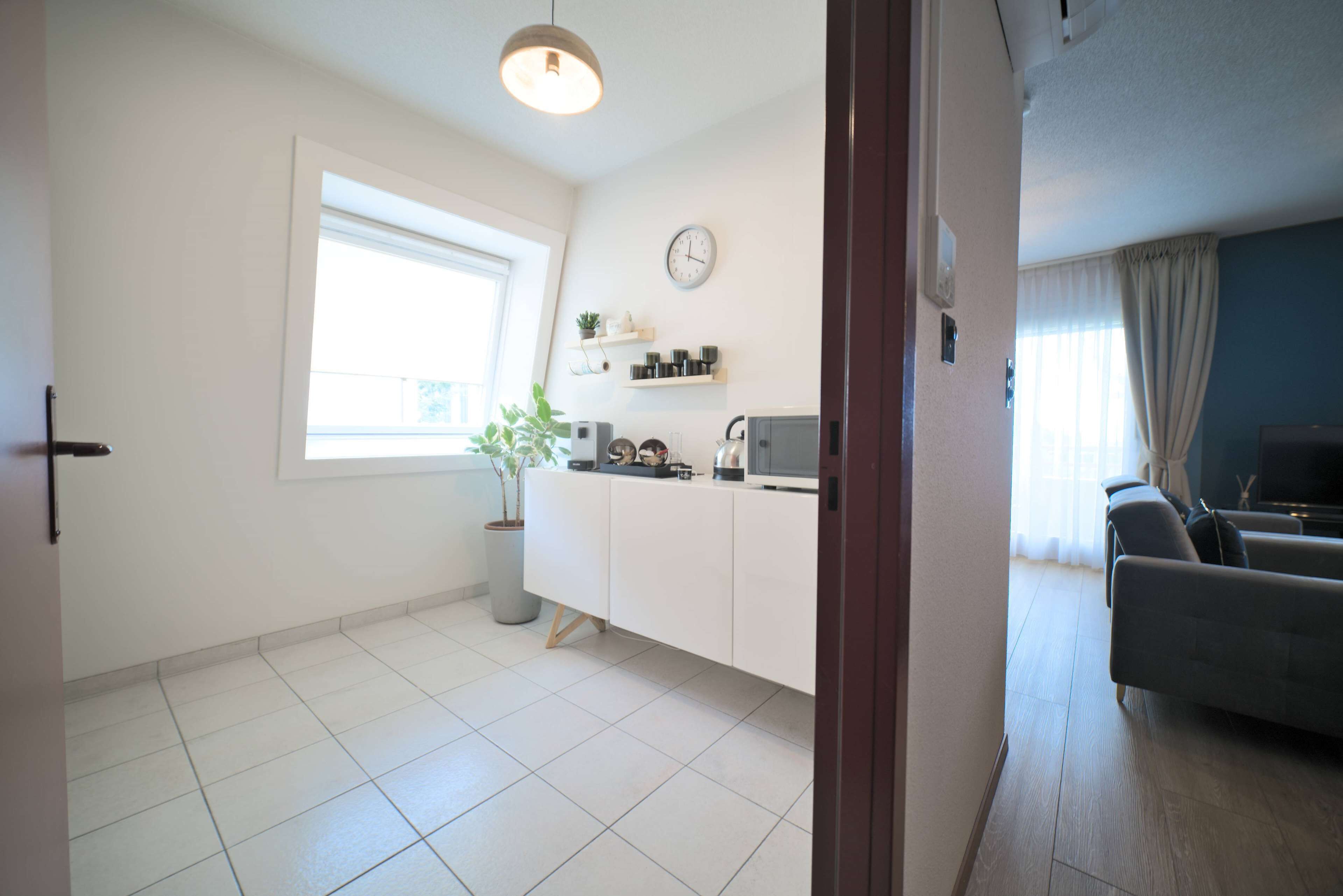 Una sala de estar moderna con un sofá gris, un televisor y una cocina con armarios blancos y una planta en maceta.