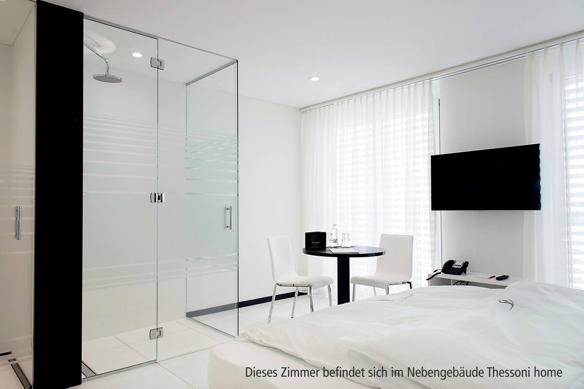 A modern hotel room with a glass shower enclosure, a bed, a small table with chairs, and a mounted TV.