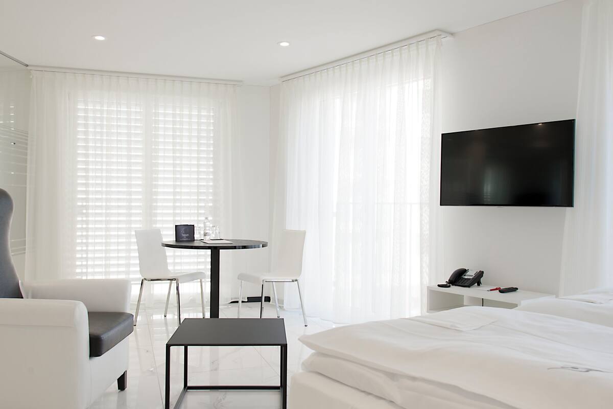 A modern hotel room with a large bed, a desk with a chair, a TV mounted on the wall, and large windows with sheer curtains.