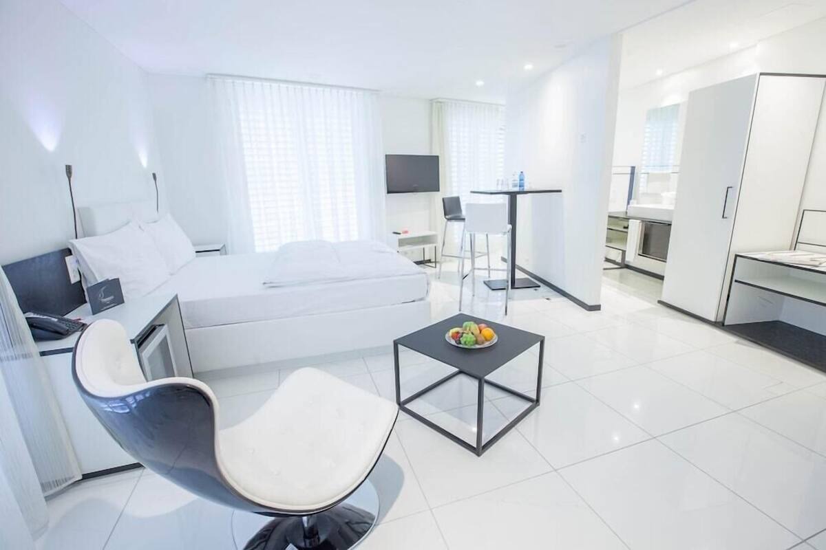 A modern hotel room with a bed, a desk, a chair, a TV, and a dining table with fruit.