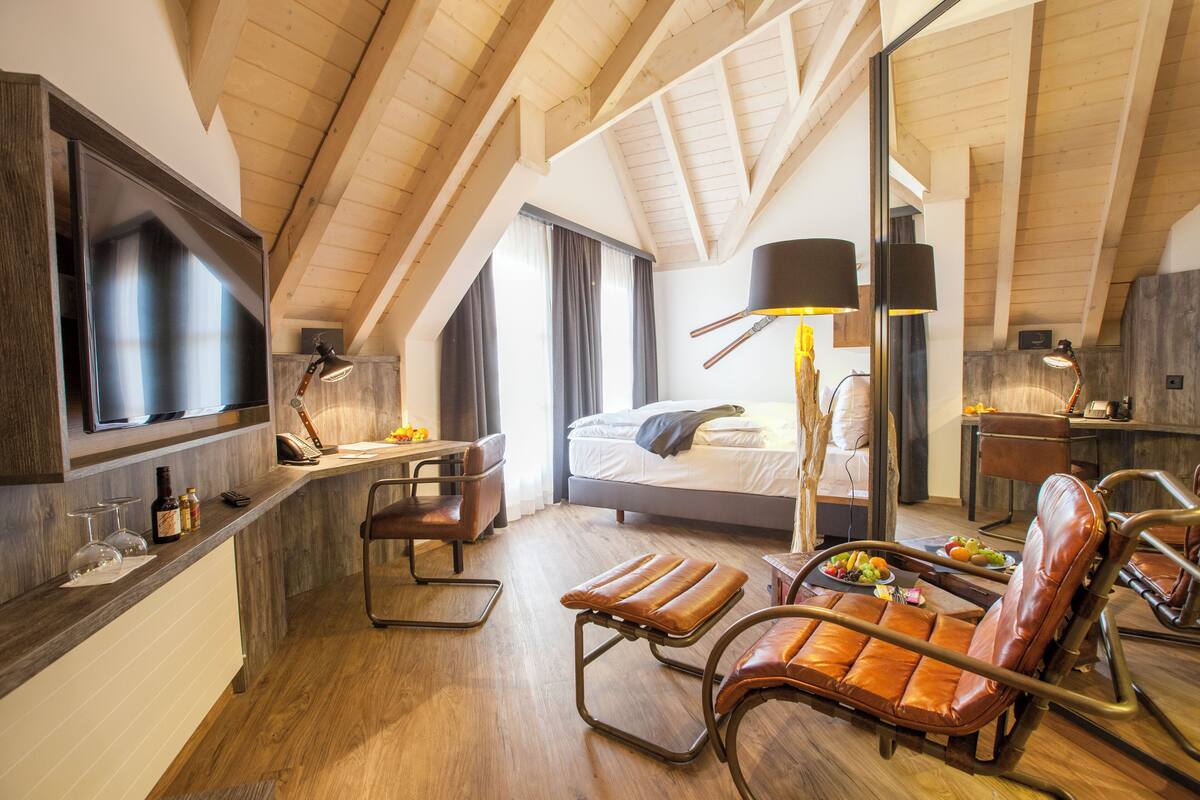 A modern hotel room with a loft-style ceiling, a bed, a desk with a chair, a dining area with a table and chairs, and a TV mounted on the wall.