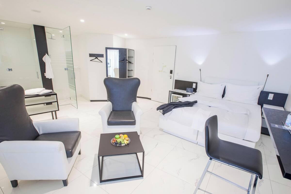 A modern hotel room with a white sofa, two armchairs, a small table with fruit, a bed with white linens, a desk with a laptop, and a glass shower area.