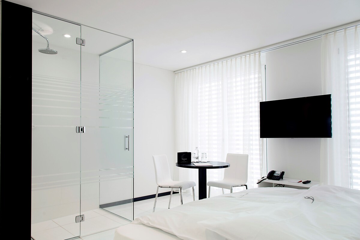A modern hotel room with a glass shower enclosure, a bed, a small table with chairs, and a mounted TV.