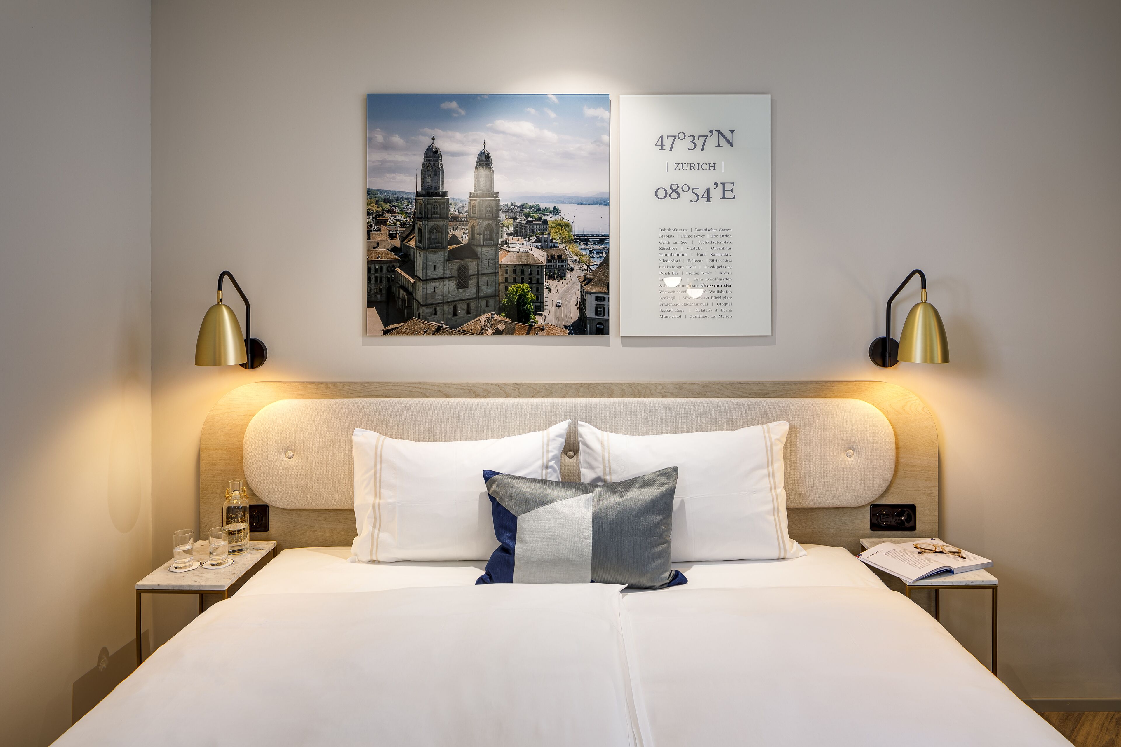 A hotel room with a large bed, two bedside tables, a nightstand, a wall-mounted lamp, a framed picture of a cityscape, and a framed map.