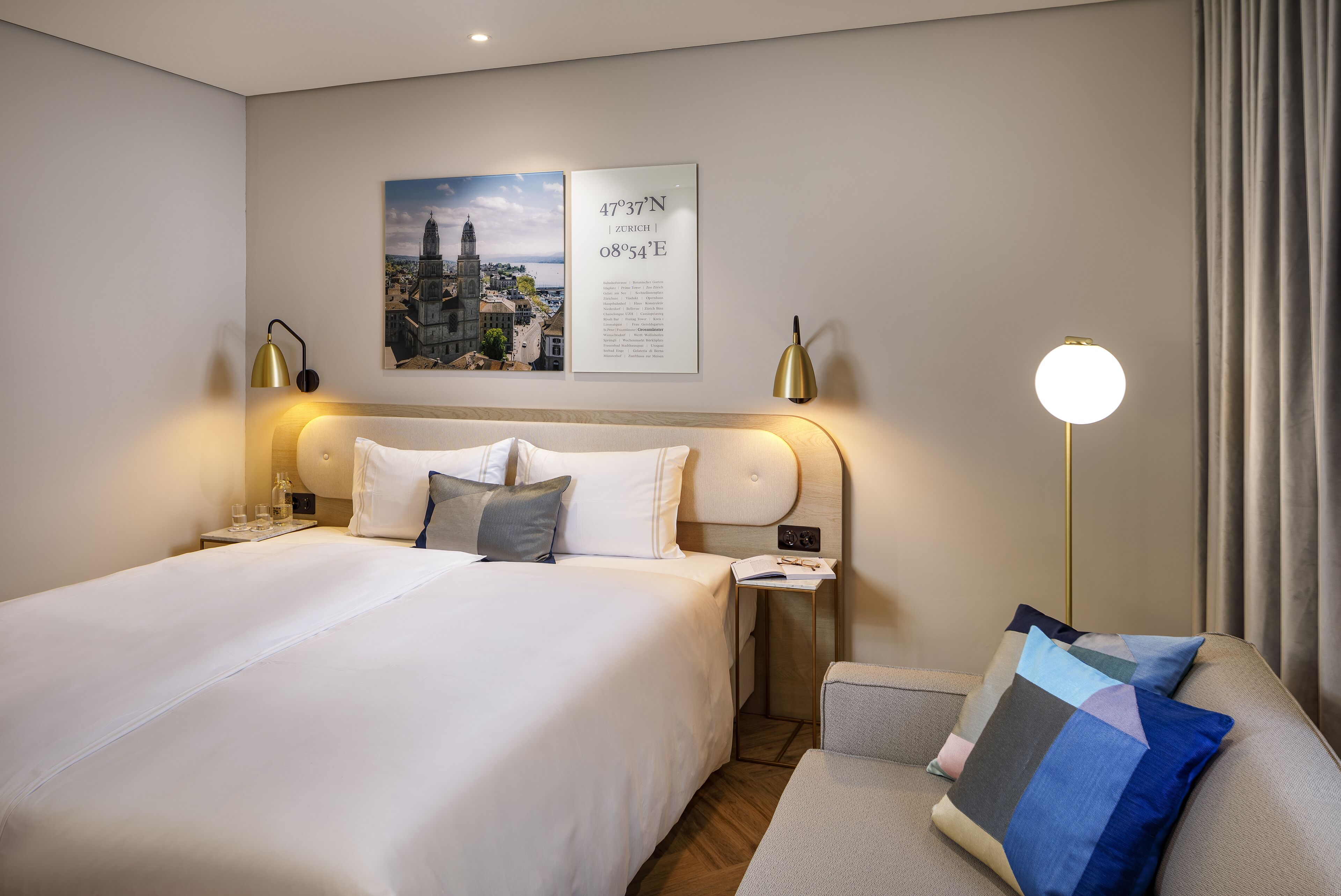 Una habitación de hotel con una cama grande, una mesita de noche con lámpara, una lámpara de lectura, una pequeña mesa con un libro y un sofá con un cojín.