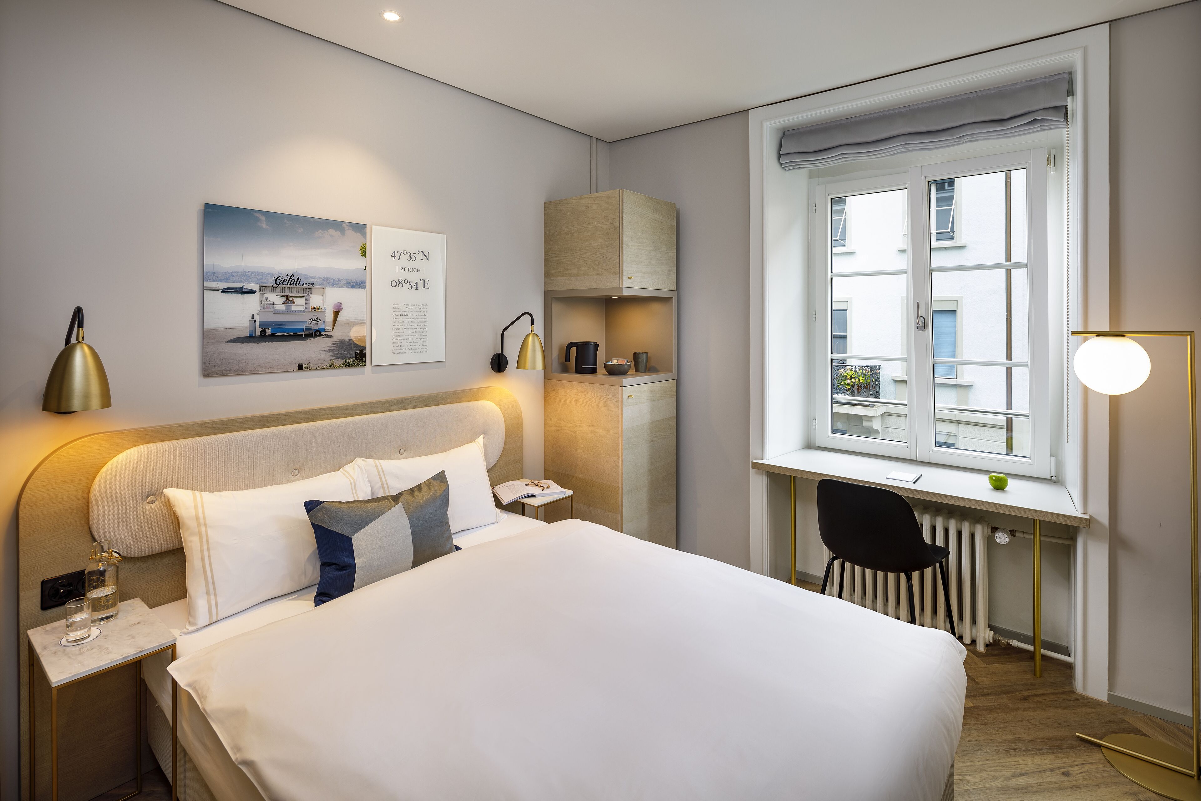 Ein modernes Hotelzimmer mit einem großen Bett, einem Schreibtisch mit Stuhl, einem Fenster mit Blick auf Gebäude und einem kleinen Tisch mit Lampe.