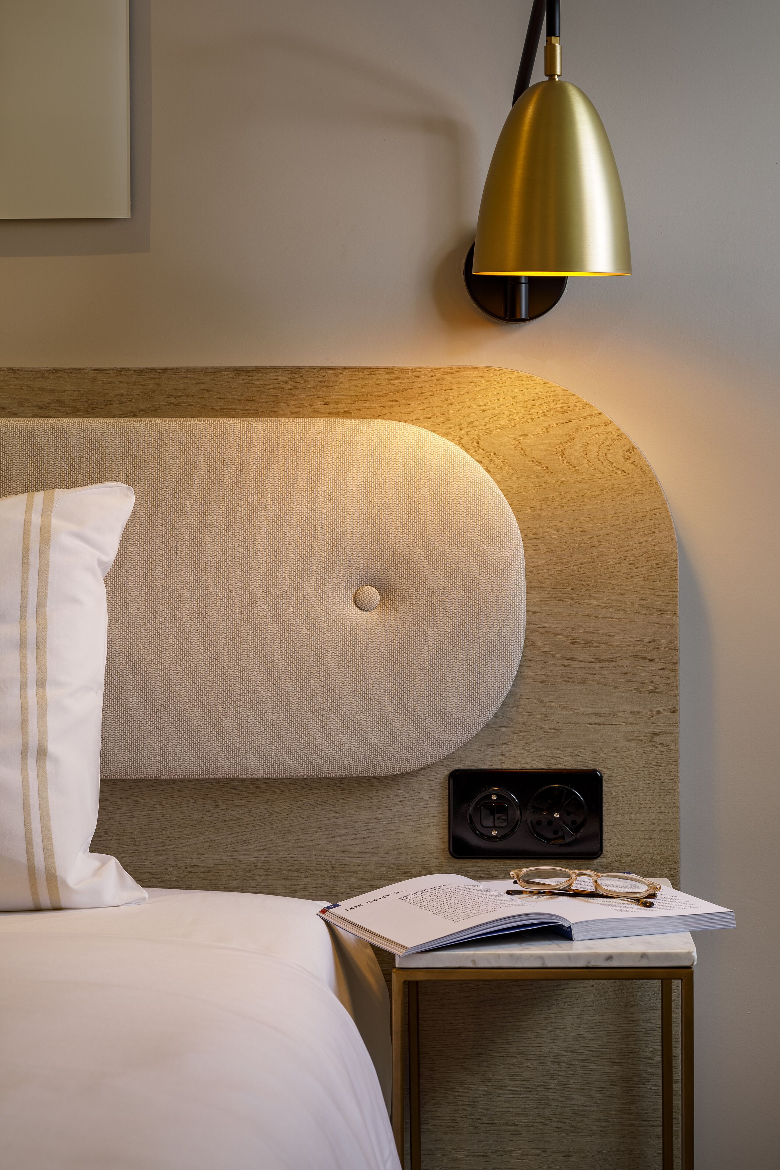 Un lit bien fait, avec une literie blanche, une petite table de chevet sur laquelle se trouvent un livre et des lunettes, et une lampe fixée au mur.