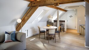 Un appartement moderne de style « loft », avec des poutres apparentes, un coin repas doté d’une table ronde et de chaises, un coin cuisine et un espace salon confortable avec un canapé et des coussins.