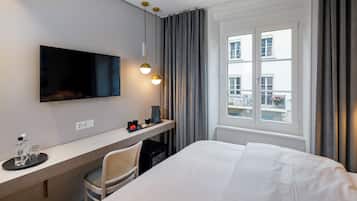 Ein Hotelzimmer mit einem Bett, einem Schreibtisch mit Fernseher, einem Sessel und einem Fenster mit Vorhängen.