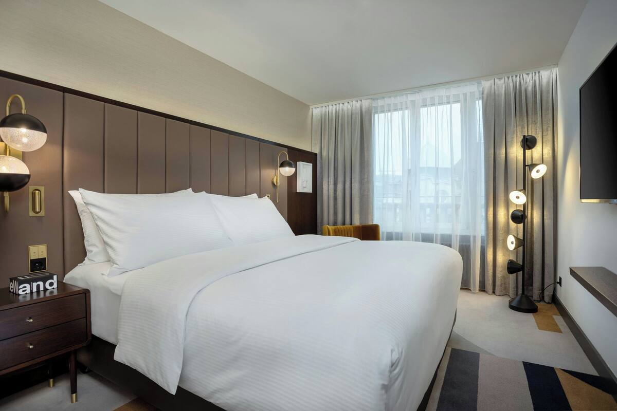 Une chambre d’hôtel avec un grand lit, une table de chevet, une lampe, une télévision et une fenêtre avec des rideaux.