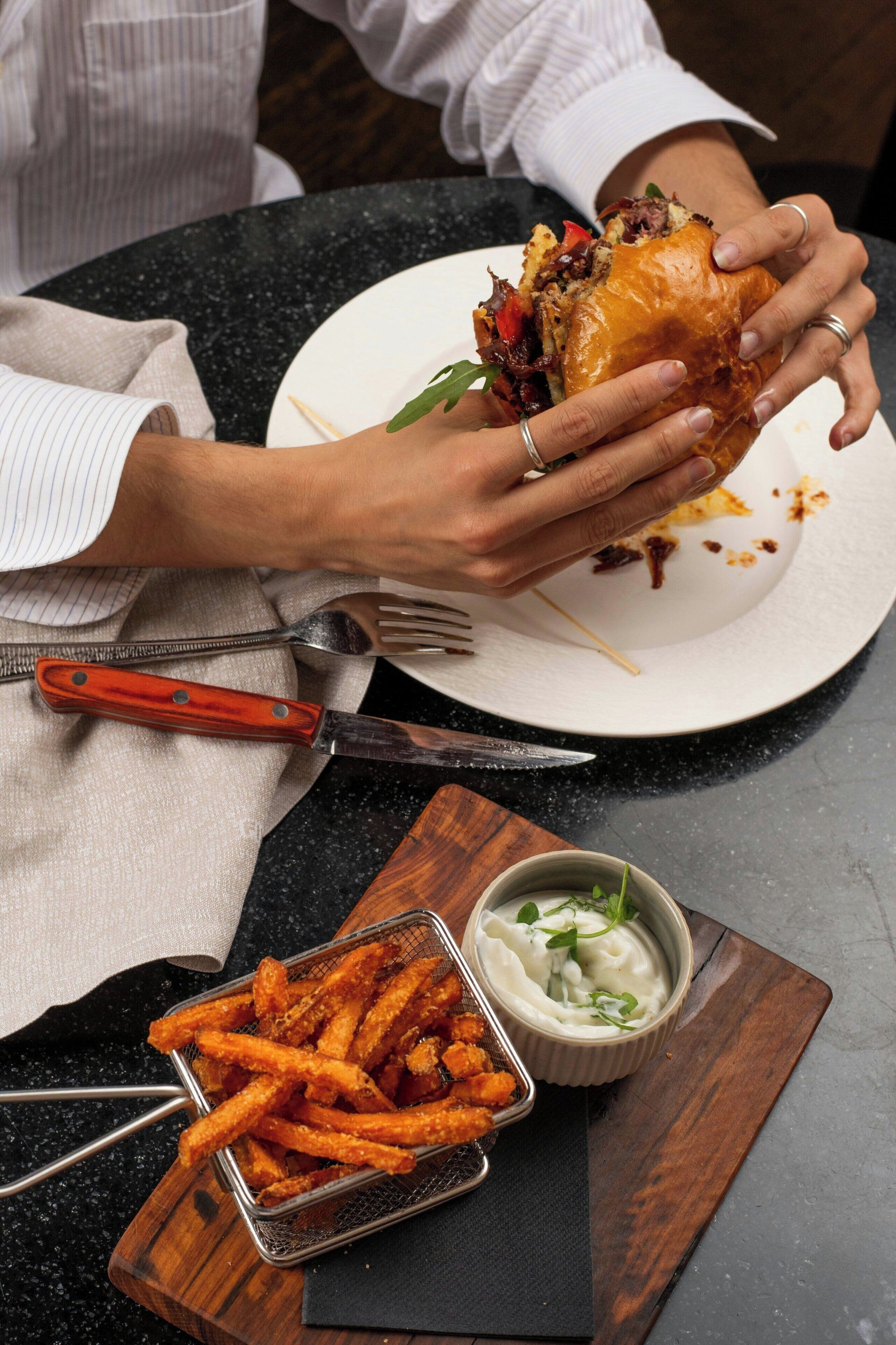 Une personne tient un sandwich à l’aide d’une fourchette et d’un couteau, et est assise à une table. Sur la table, on peut voir un bol de sauce et un panier de frites.