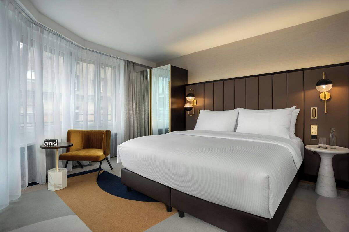 Une chambre d’hôtel moderne avec un grand lit, une table de chevet, une chaise et une vue sur la ville à travers de fines rideaux.