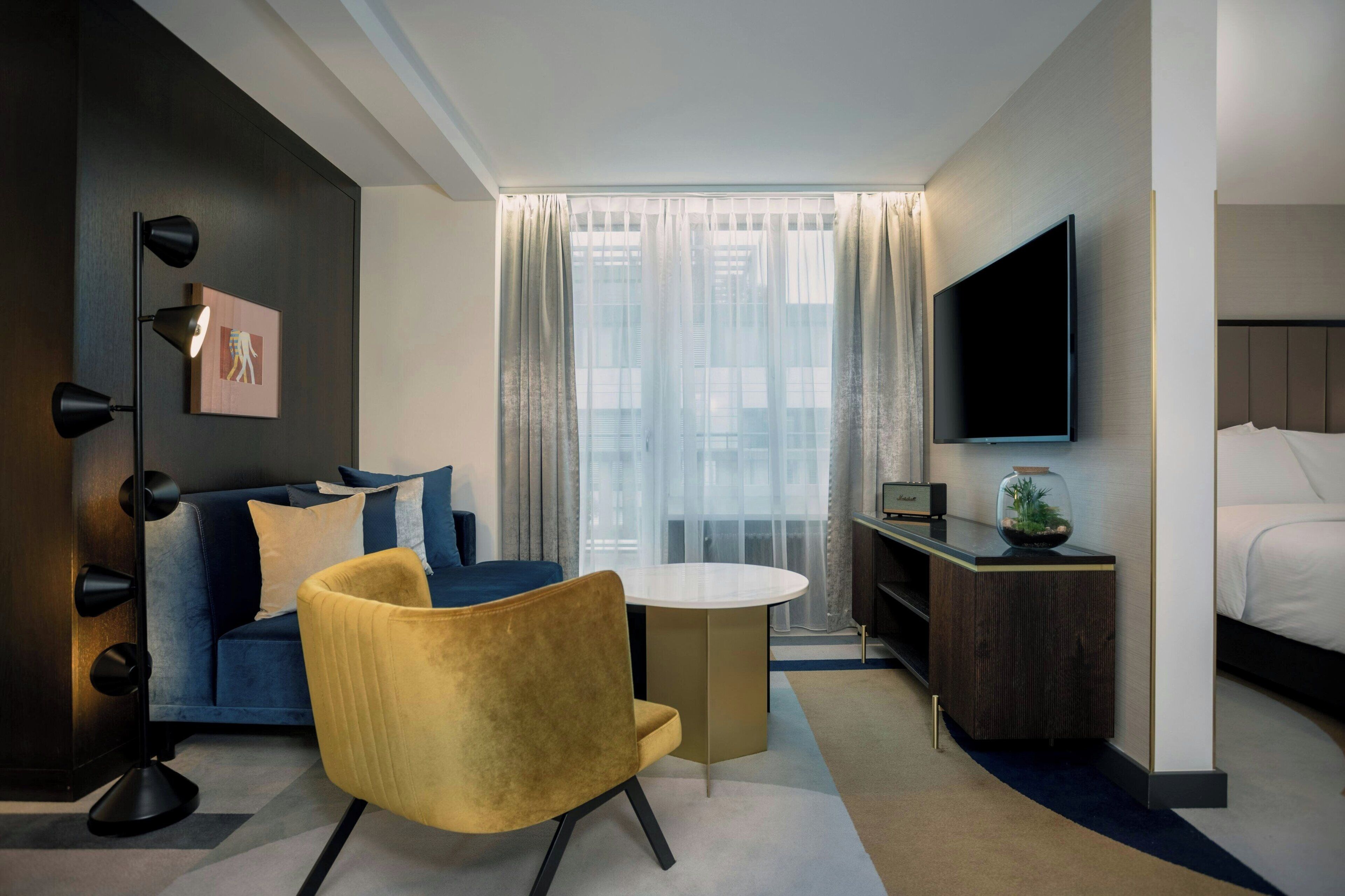 Ein modernes Hotelzimmer mit Sofa, gelbem Sessel, rundem Tisch, Fernseher und Bett.