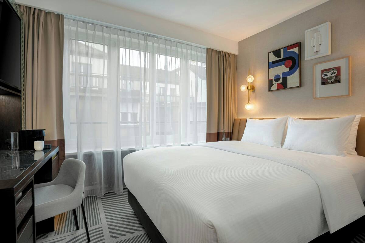 Une chambre d’hôtel avec un grand lit, un bureau avec une chaise, une télévision et une fenêtre avec des rideaux légers.