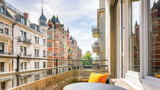 Een balkon met een ronde tafel en een geel kussen, met uitzicht op een rij historische gebouwen met versierde architectuur.