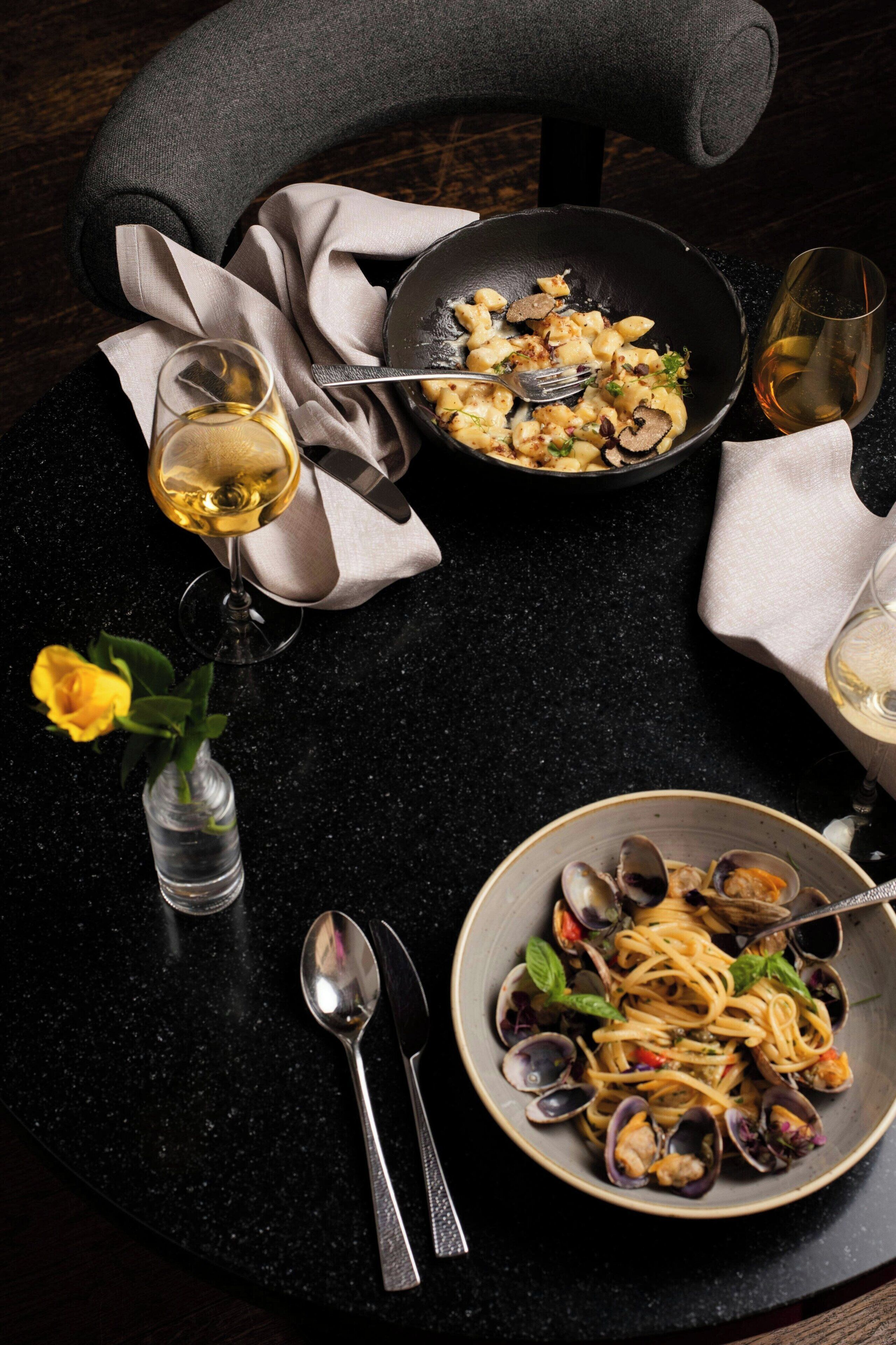 A dining table with two plates of pasta, a bowl of clams, a wine glass, and a vase with a yellow rose.