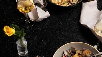 Een eettafel met twee borden pasta, een kom met vongole, een wijnglas en een vaas met een gele roos.