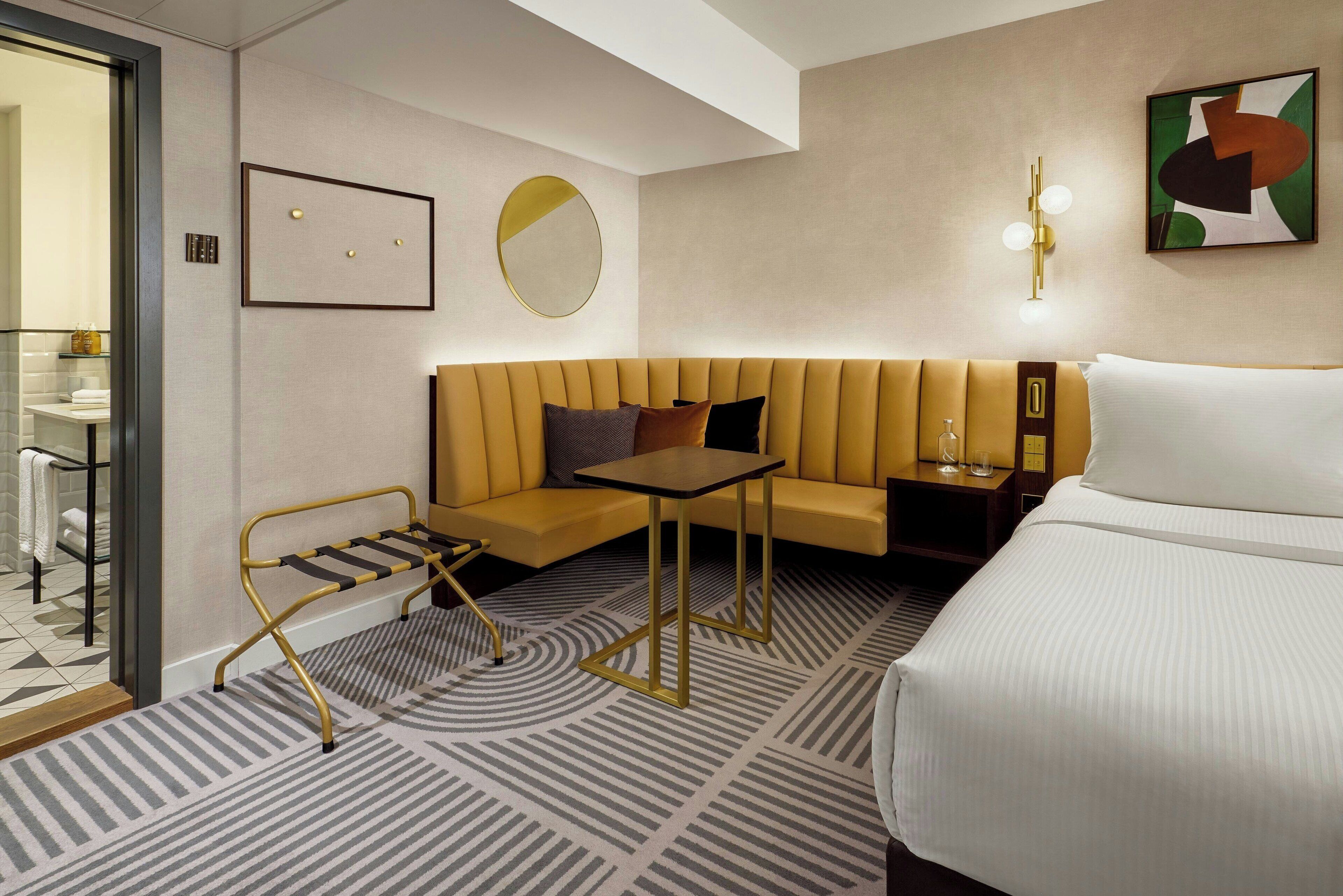 A hotel room with a bed, a sofa, a small table, a bench, a mirror, and a wall-mounted light fixture.
